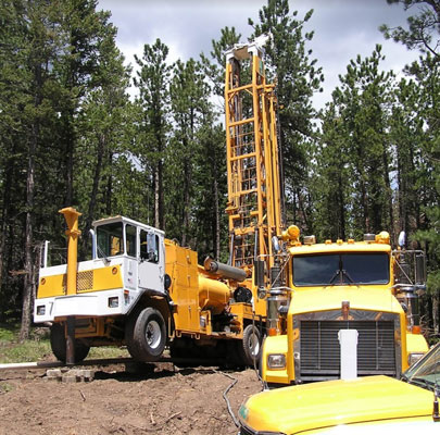 A large orange and white well drilling rig with derrick extending upwards toward the towering treeline behind it